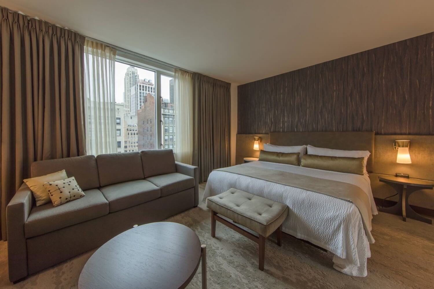 Guestroom with king bed, white linens, gray couch, gold accents, and city views