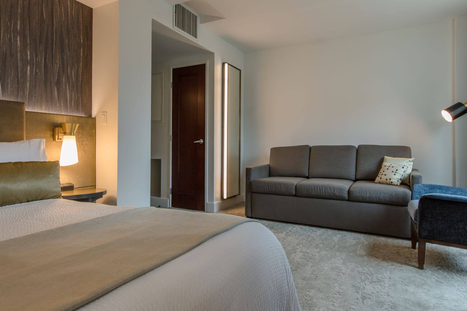 Guestroom with king bed, white linens, gray couch, gold accents, and a blue chair